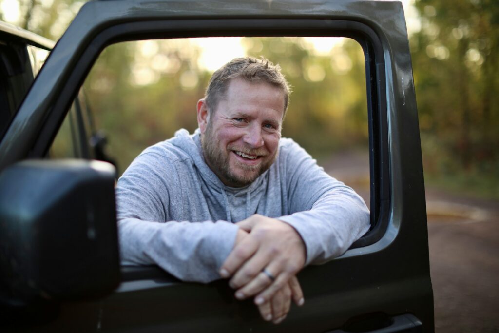 Un homme d'une quarantaine d'année souriant appué sur le rebord de fenêtre d'une camionette.