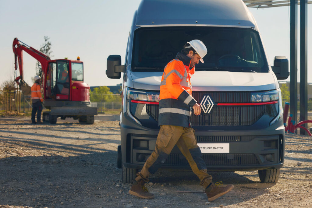 Un ouvrier sur un chantier passe devant un fourgon Renault Trucks Master, et on voit un tracteur en arrière-plan.